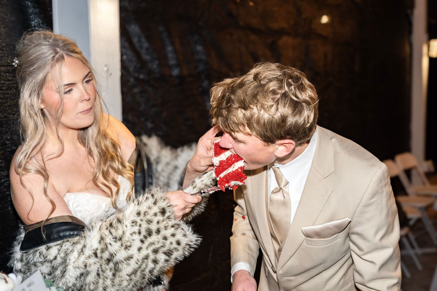 Groom in tan suit playfully eating red velvet cake while bride in fur wrap watches at reception.