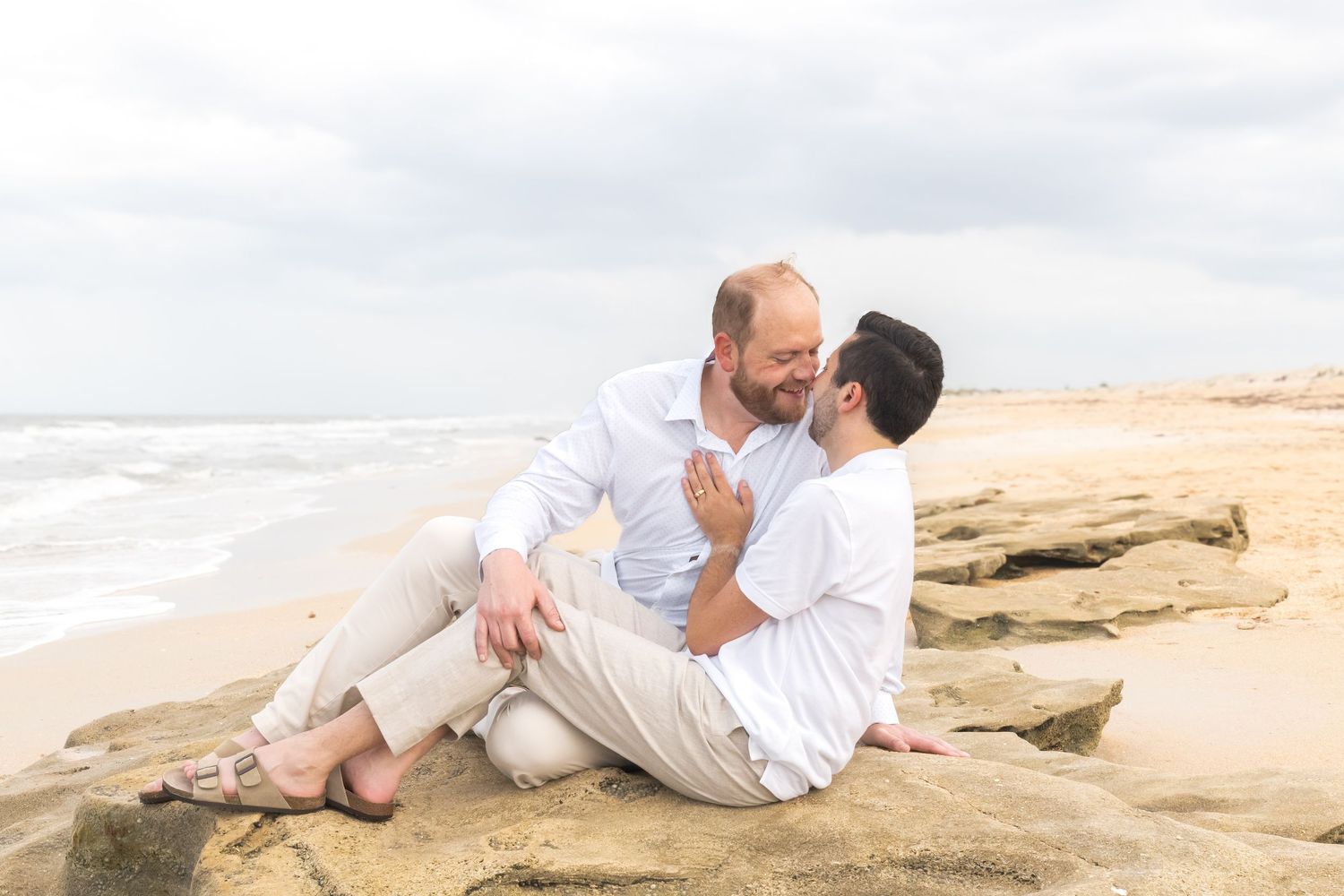 Two men sitting on beach rocks, sharing a romantic moment by the ocean on a cloudy day.