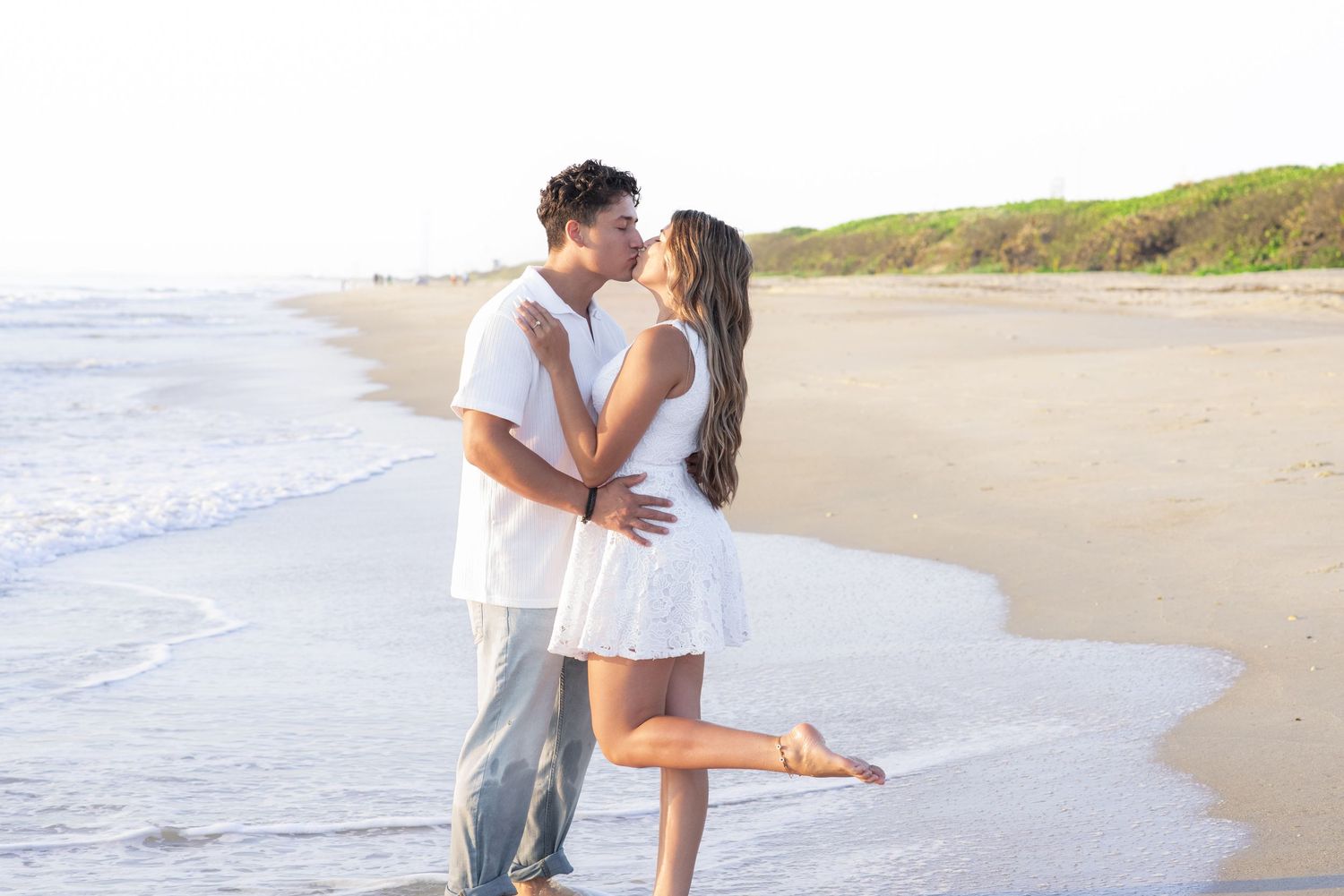 Romantic couple sharing a kiss on a sandy beach, waves gently lapping the shore, dressed in white outfits.