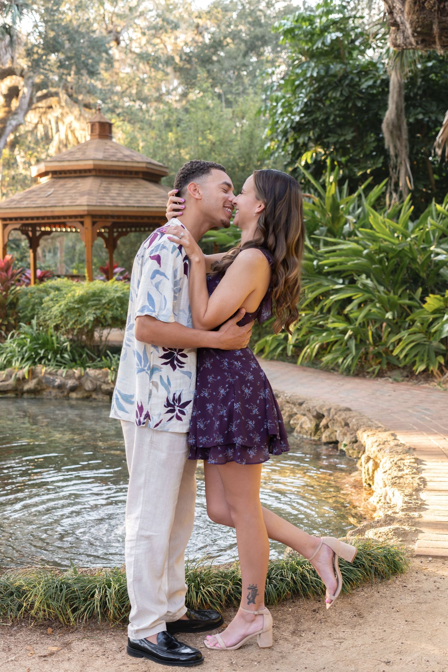 Couple sharing a romantic kiss by a tranquil garden pond with a gazebo and lush greenery backdrop.