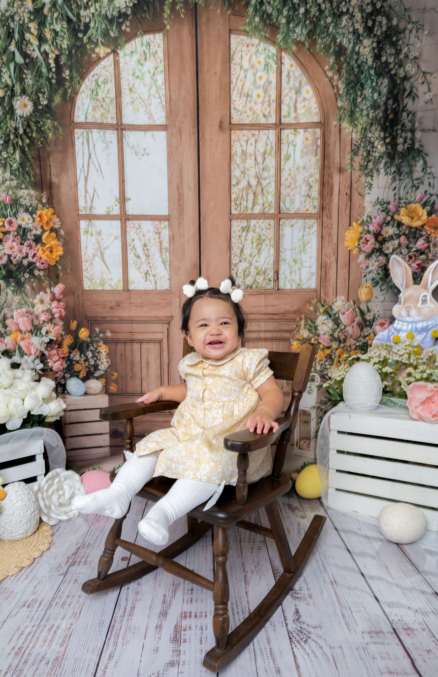 Happy baby girl in yellow dress sitting in a rocking chair with an Easter-themed floral backdrop and decorations.