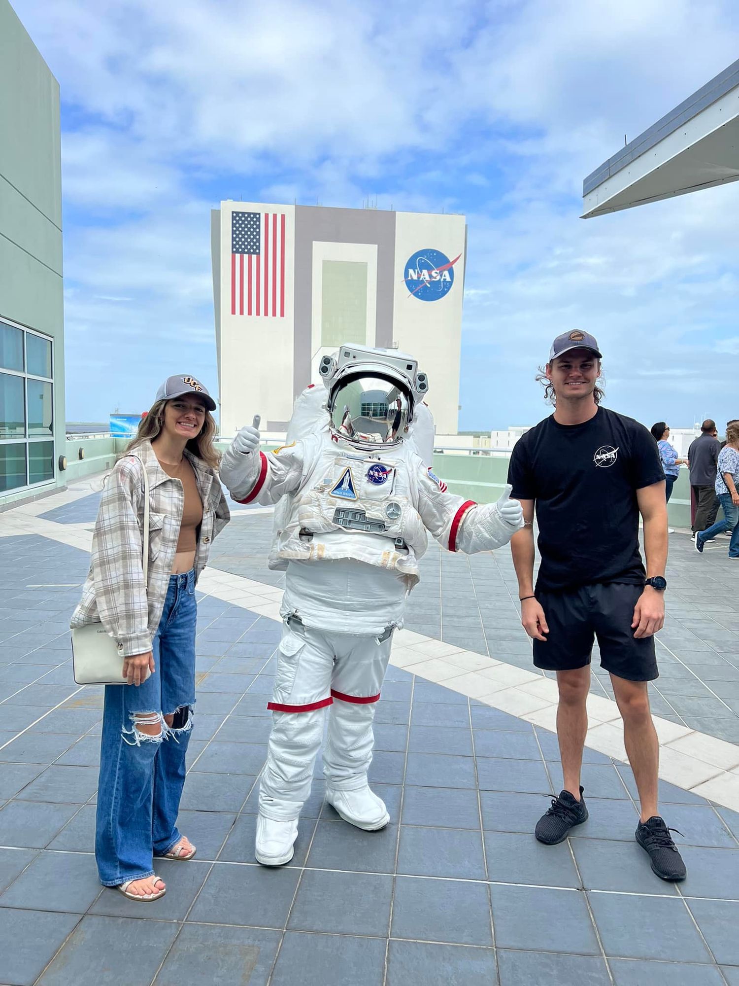Two people posing with a NASA astronaut suit mascot outside the Kennedy Space Center Vehicle Assembly Building.