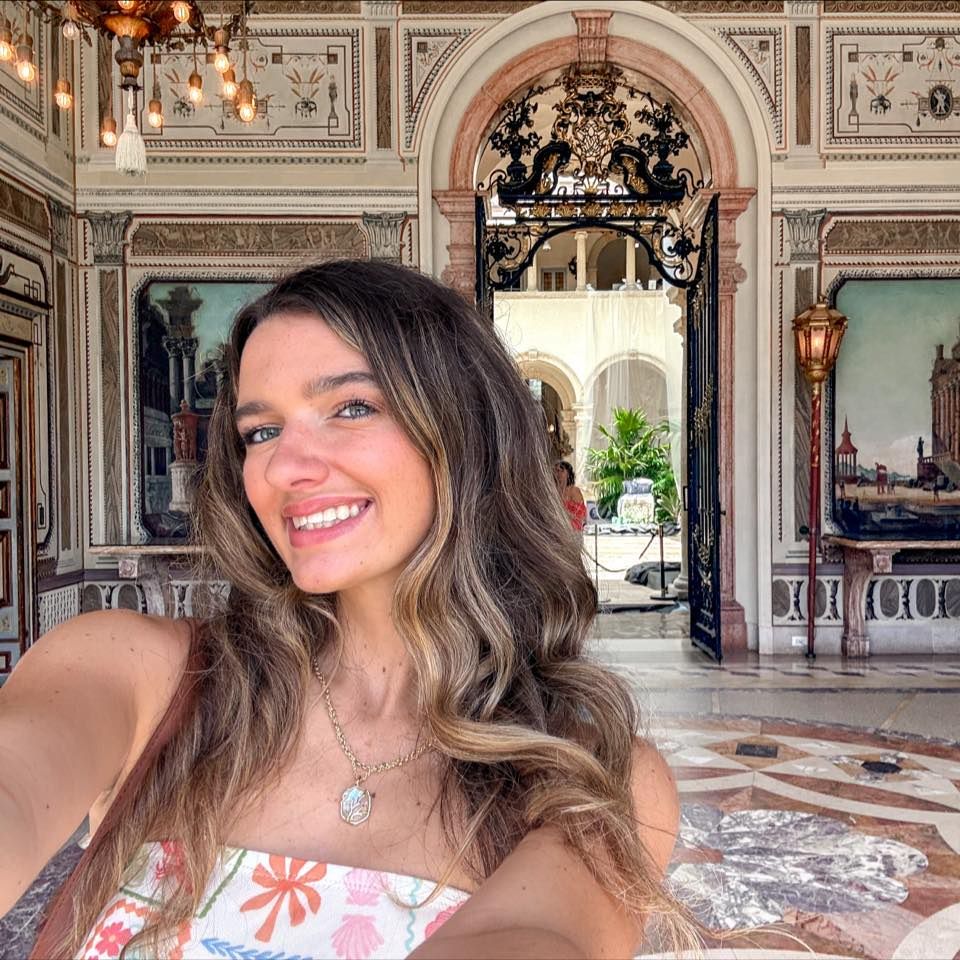 Smiling woman taking selfie in opulent gilded room with arched doorways and decorative mural paintings.
