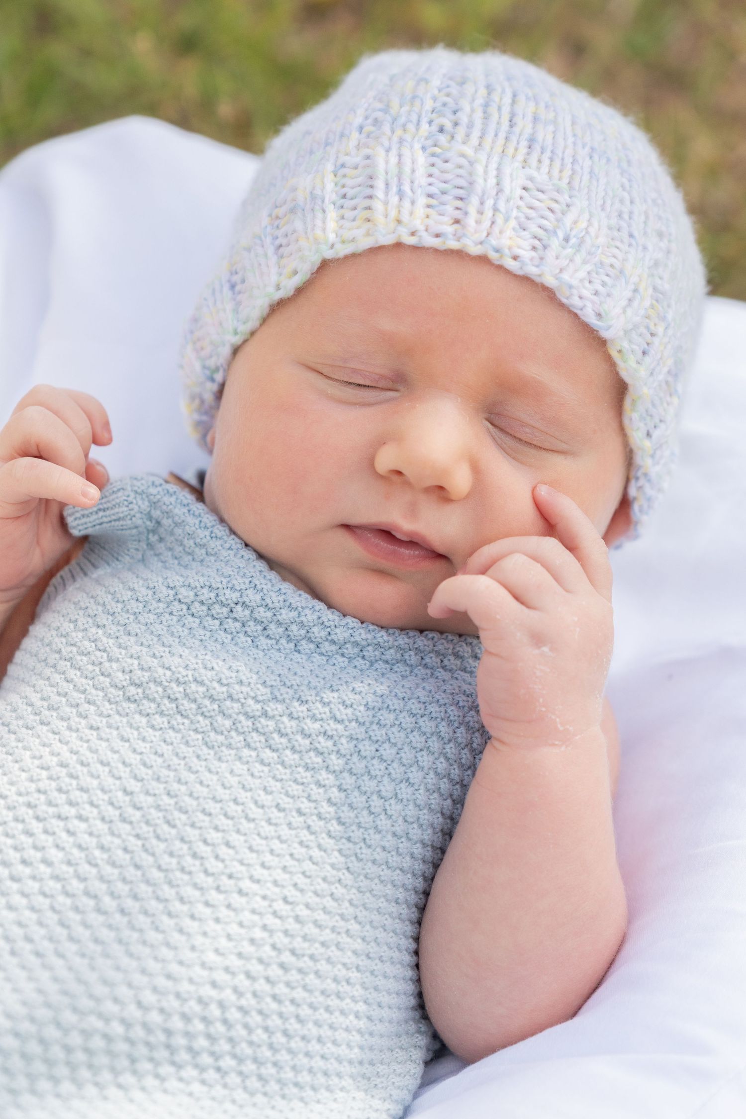 Sleeping newborn baby wearing a light blue knit hat and matching blanket, with tiny hands near face.
