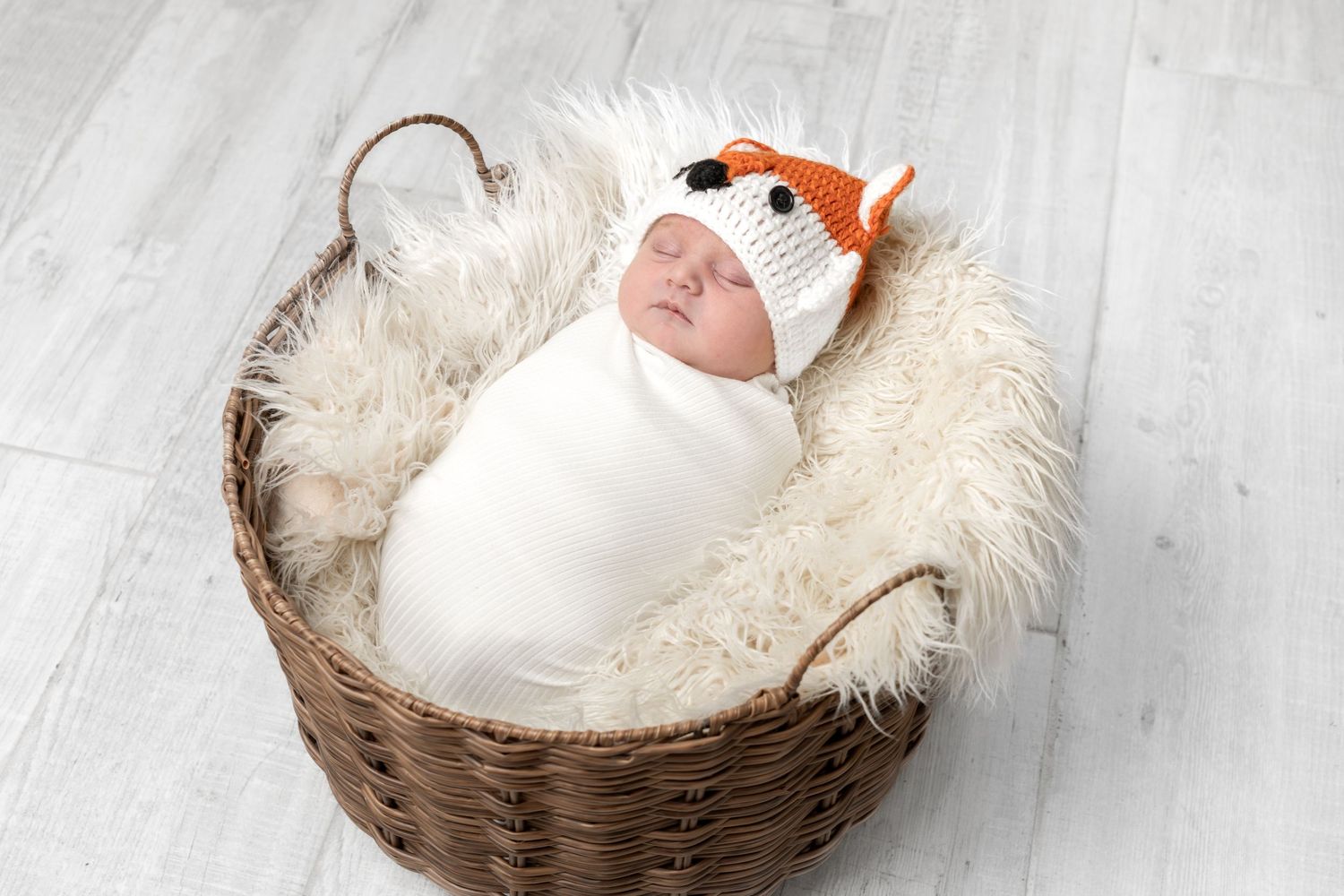 Newborn baby swaddled in white wrap, wearing a crochet fox hat, sleeping in a wicker basket lined with fluffy fur.