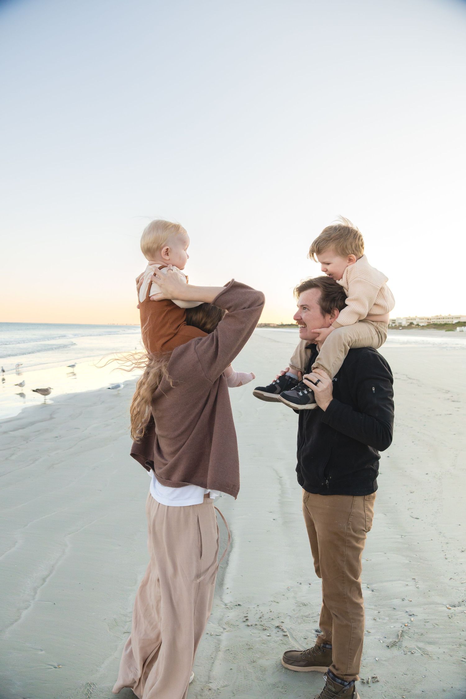 Family of four enjoying a sunset beach walk, with parents carrying young children on their shoulders.