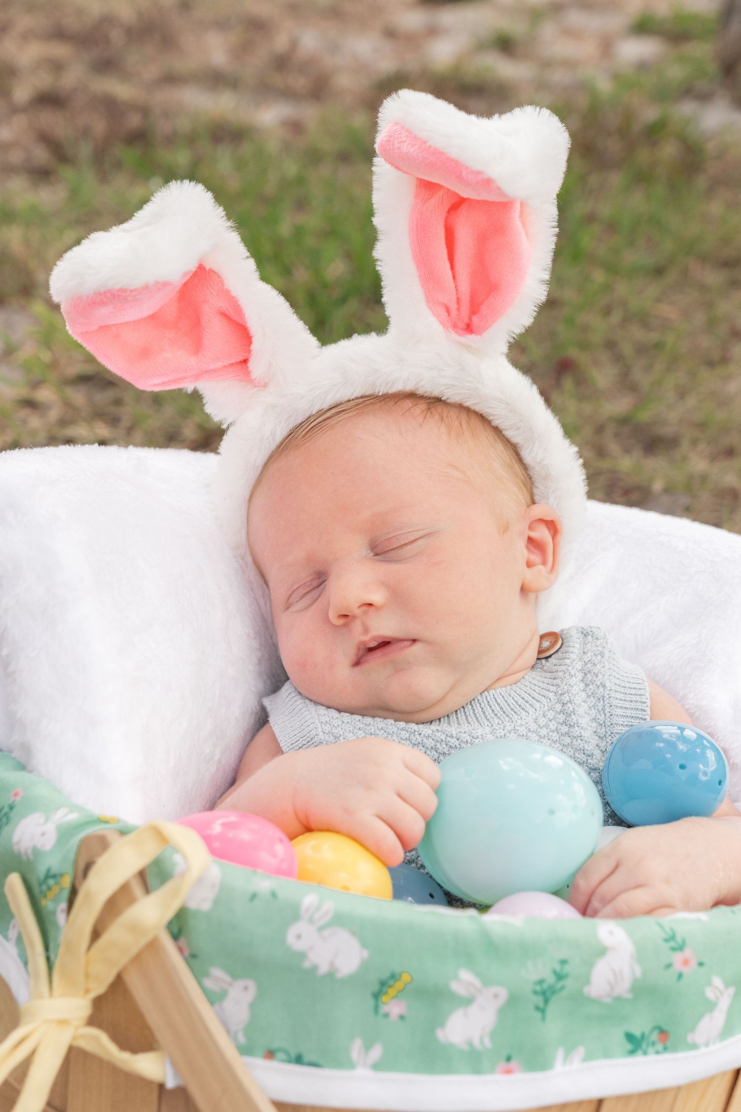 Sleeping newborn baby wearing white bunny ears headband nestled in Easter basket with colorful pastel eggs outdoors.