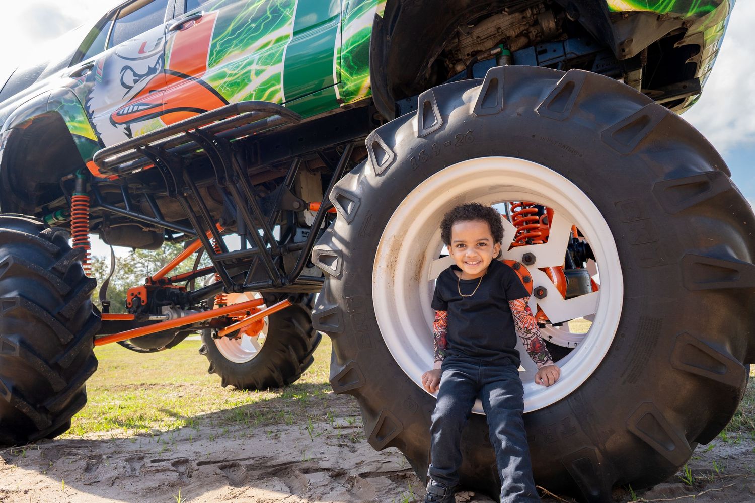 Young child smiling beside a massive monster truck tire outdoors, showcasing the vehicle's enormous scale.