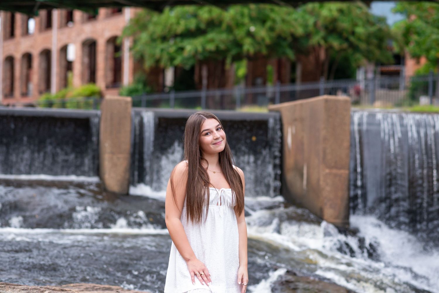 Young woman in white dress posing near a waterfall feature with brick building backdrop.