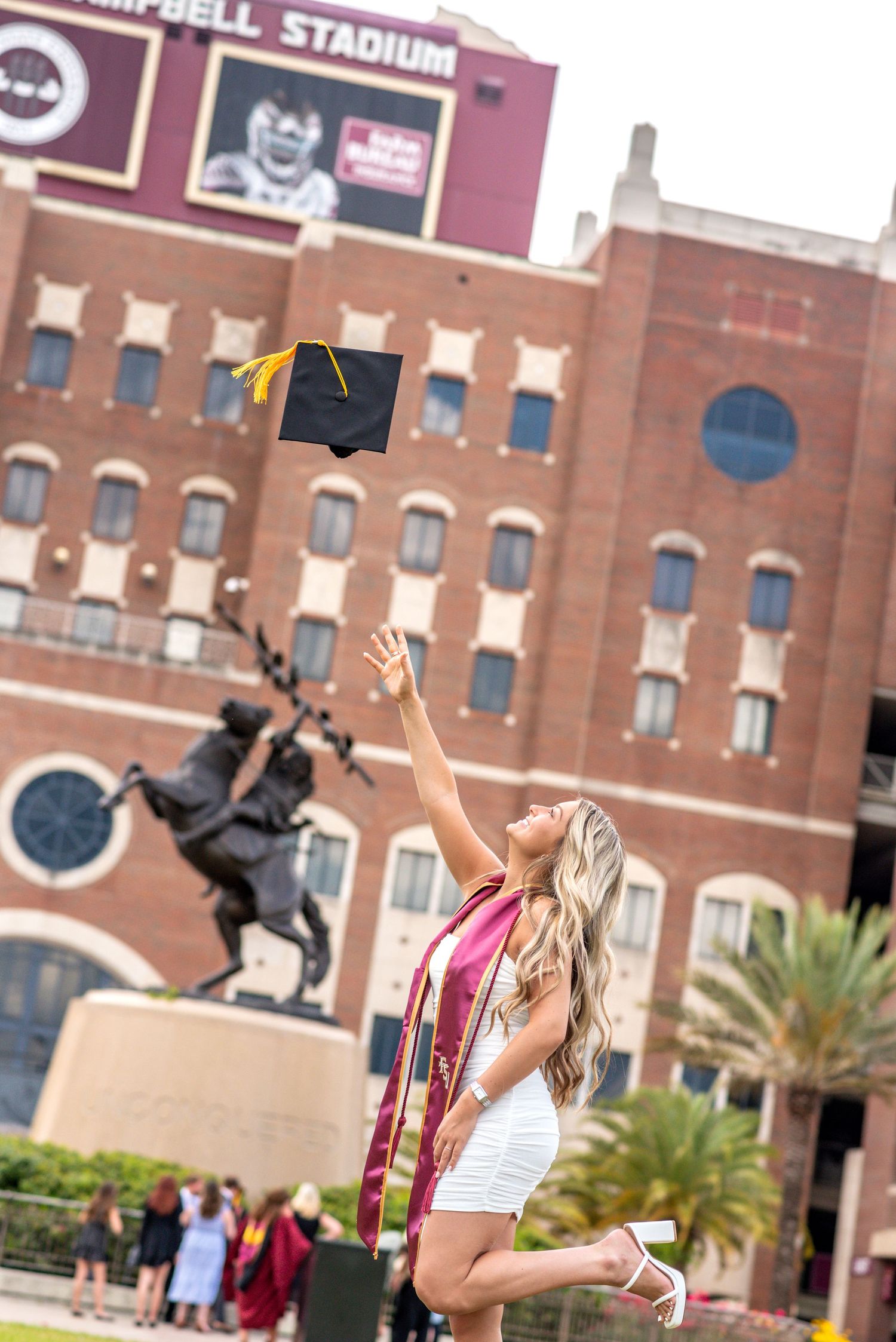 FSU graduate tosses mortarboard cap in front of Doak Campbell Stadium with Unconquered statue backdrop.