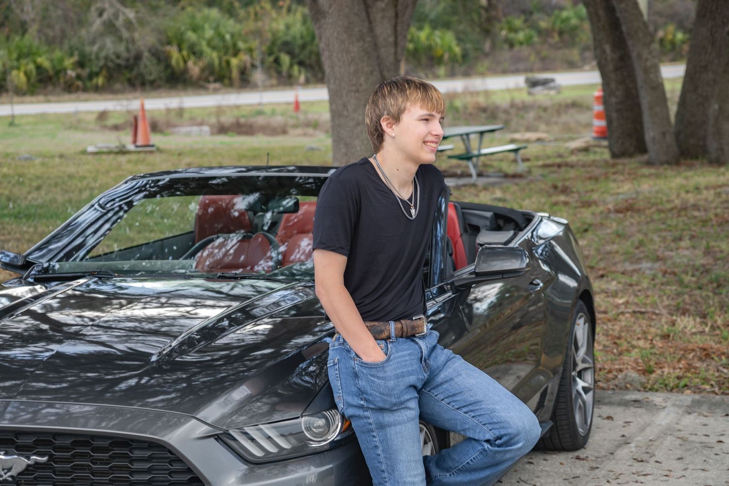 Teenage boy in black t-shirt and jeans leaning casually against a gray convertible Ford Mustang outdoors.