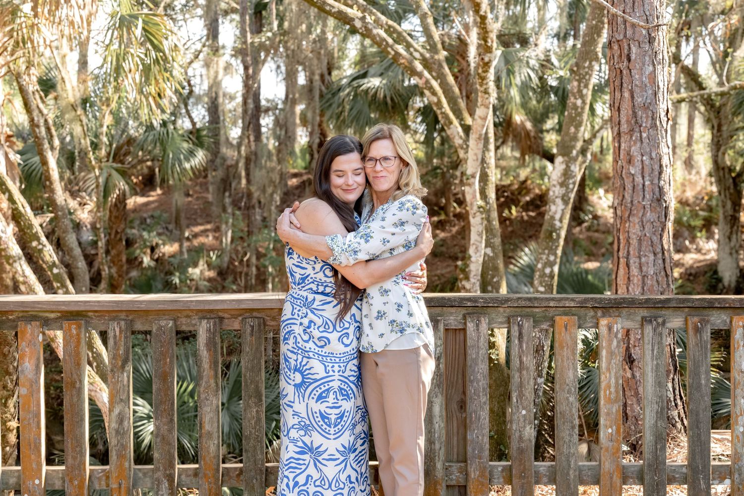 Two women hugging and smiling on a wooden deck surrounded by lush green trees in an outdoor setting.