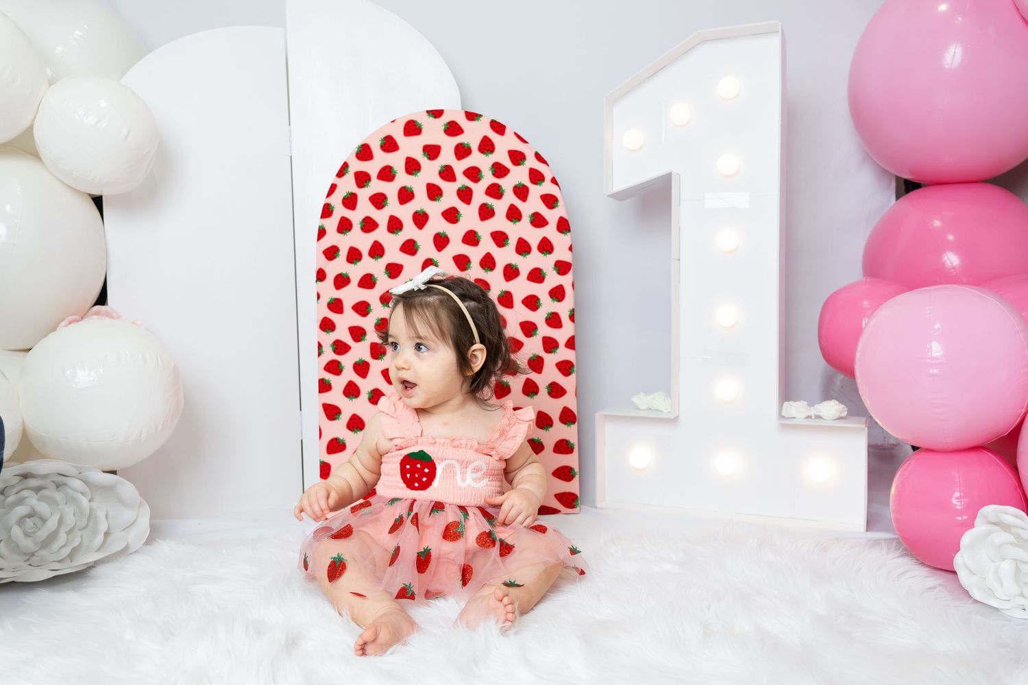 Baby girl in strawberry dress sits before a large number 1 sign with pink balloons for her first birthday.