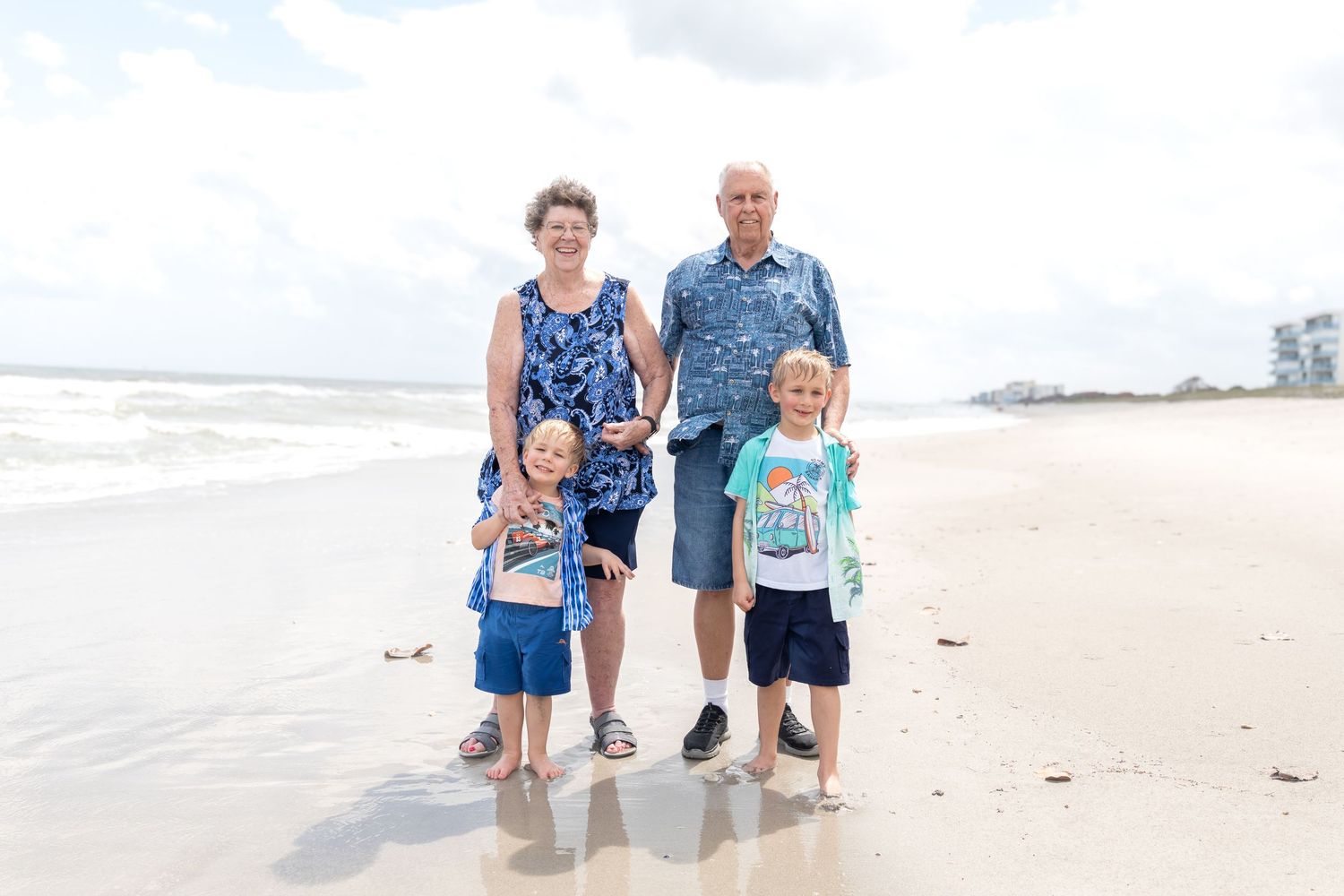Family of four posing on a sandy beach, two grandparents holding hands with two young boys near the ocean waves.
