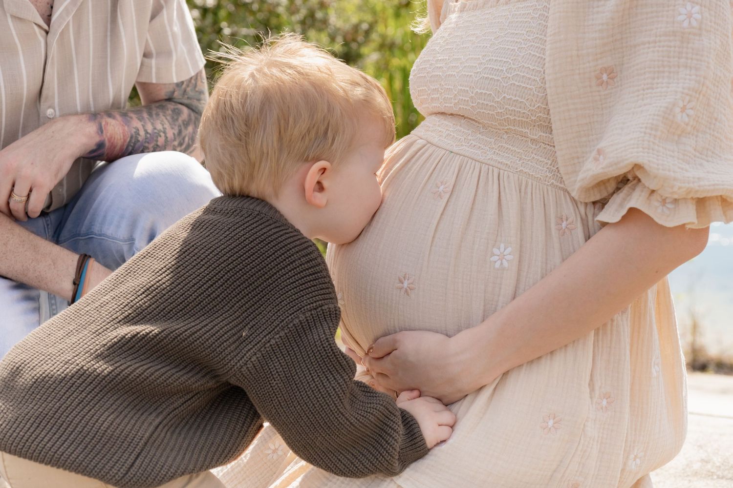 Young toddler boy kissing pregnant mother's belly during outdoor family maternity photo session.