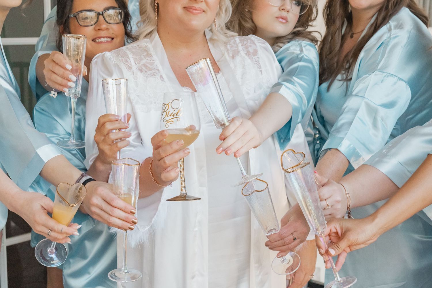 Bride and bridesmaids in light blue satin robes toast with champagne glasses during wedding preparations.