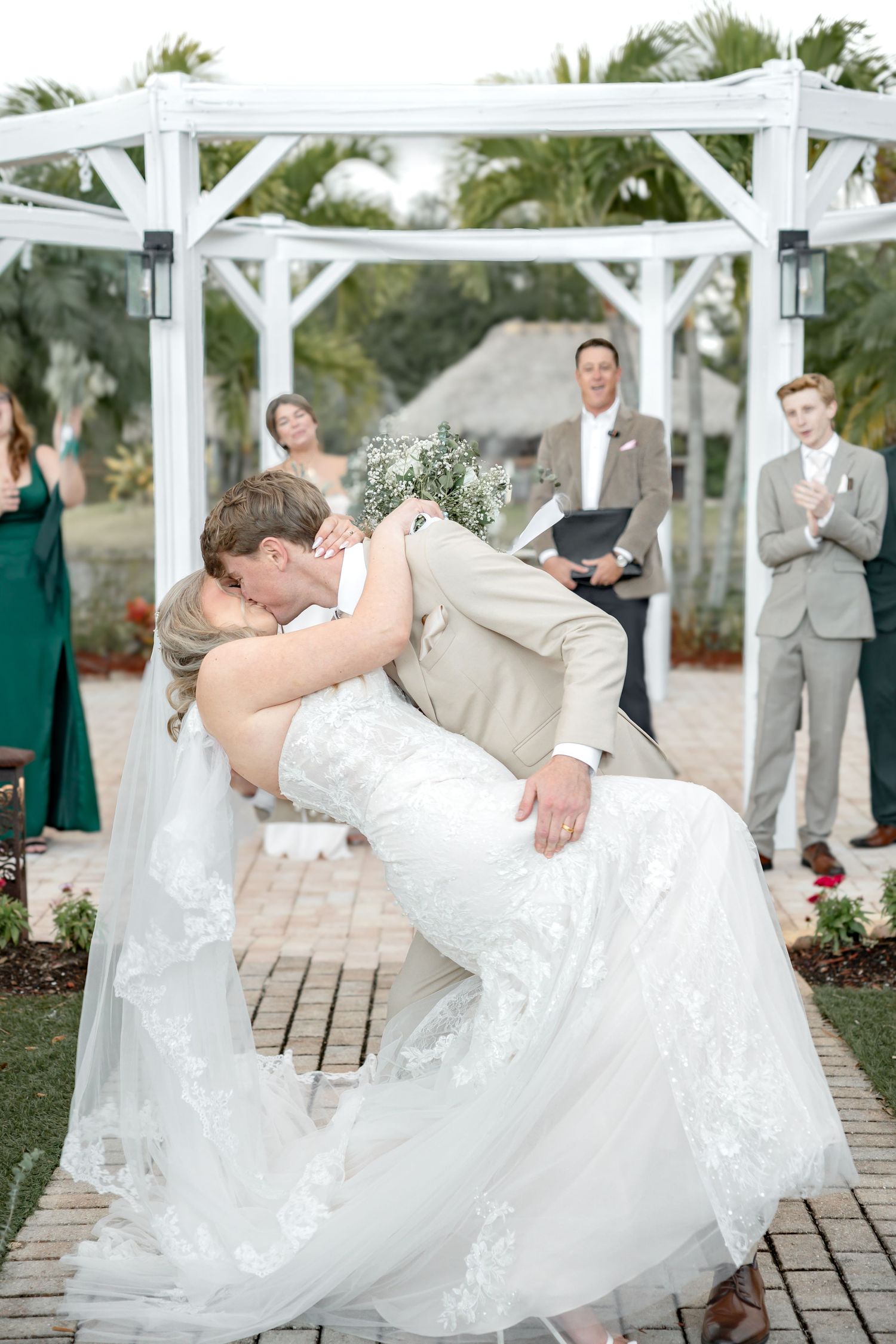 Romantic dip pose during wedding ceremony under white pergola with palm trees in background.