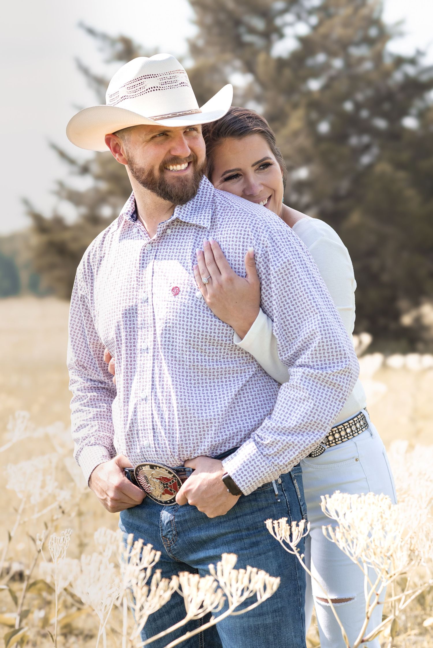A smiling couple in Western attire poses in a golden field, man in cowboy hat and plaid shirt, woman embracing him from behind.