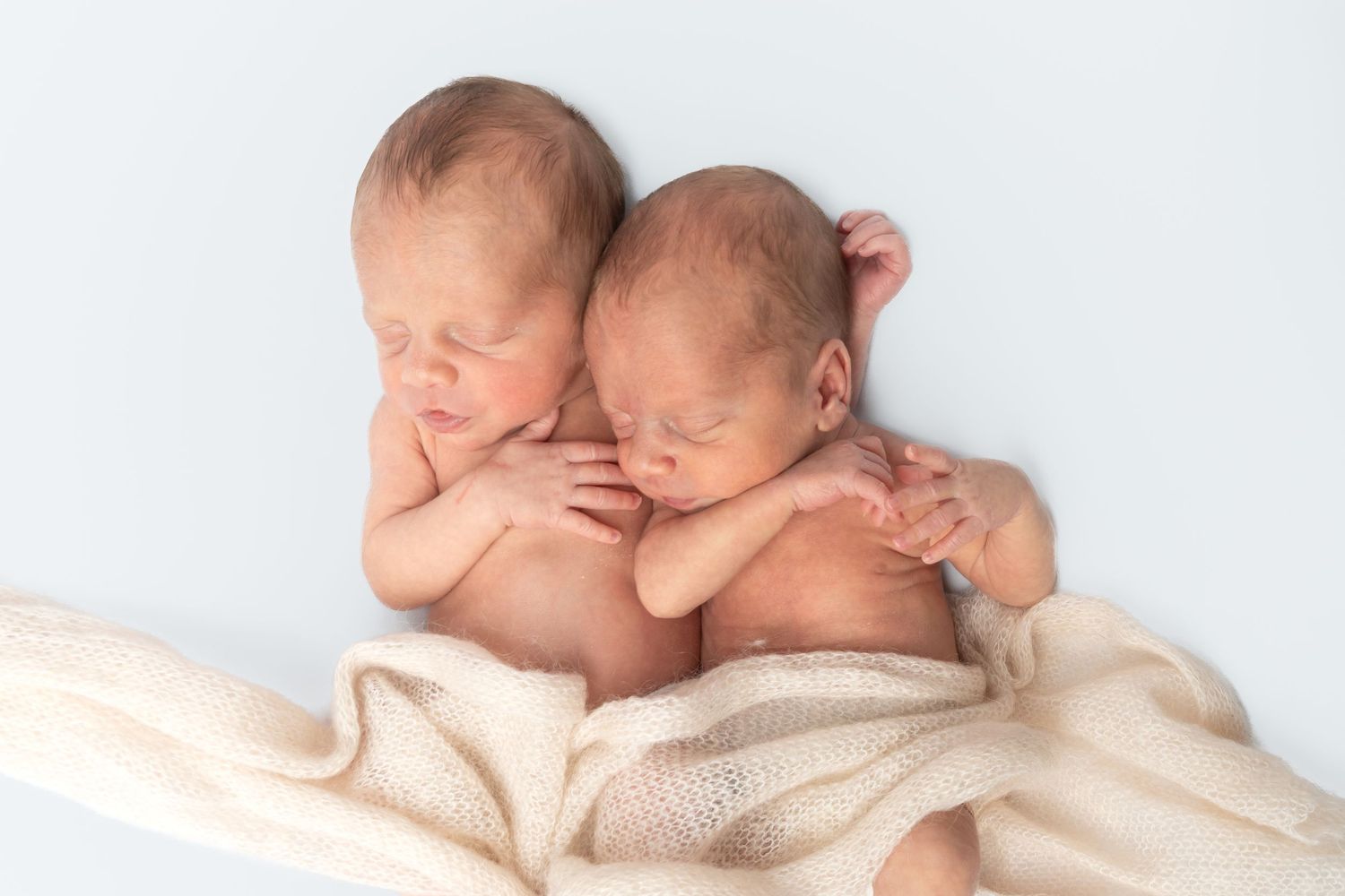 Two newborn twins cuddling together wrapped in a cream blanket on a light blue background.