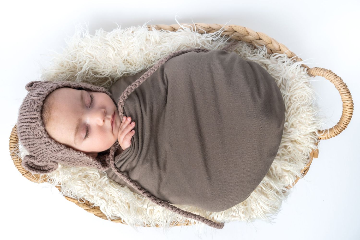 Newborn baby swaddled in brown wrap wearing knit bear bonnet, sleeping peacefully in wicker basket with fluffy white fur.