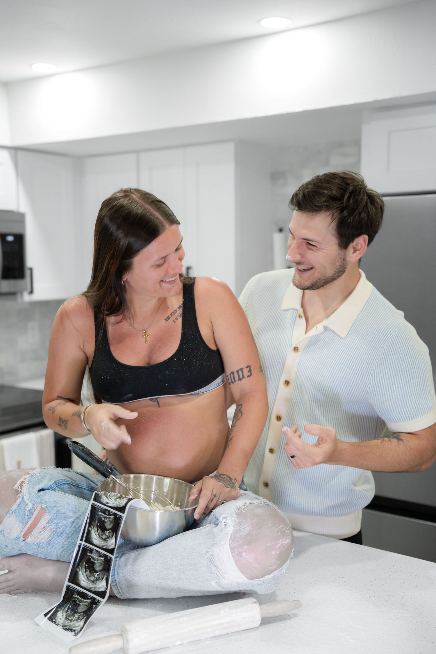 Pregnant woman holding a pot on her belly with partner, reviewing ultrasound photos in a modern kitchen.