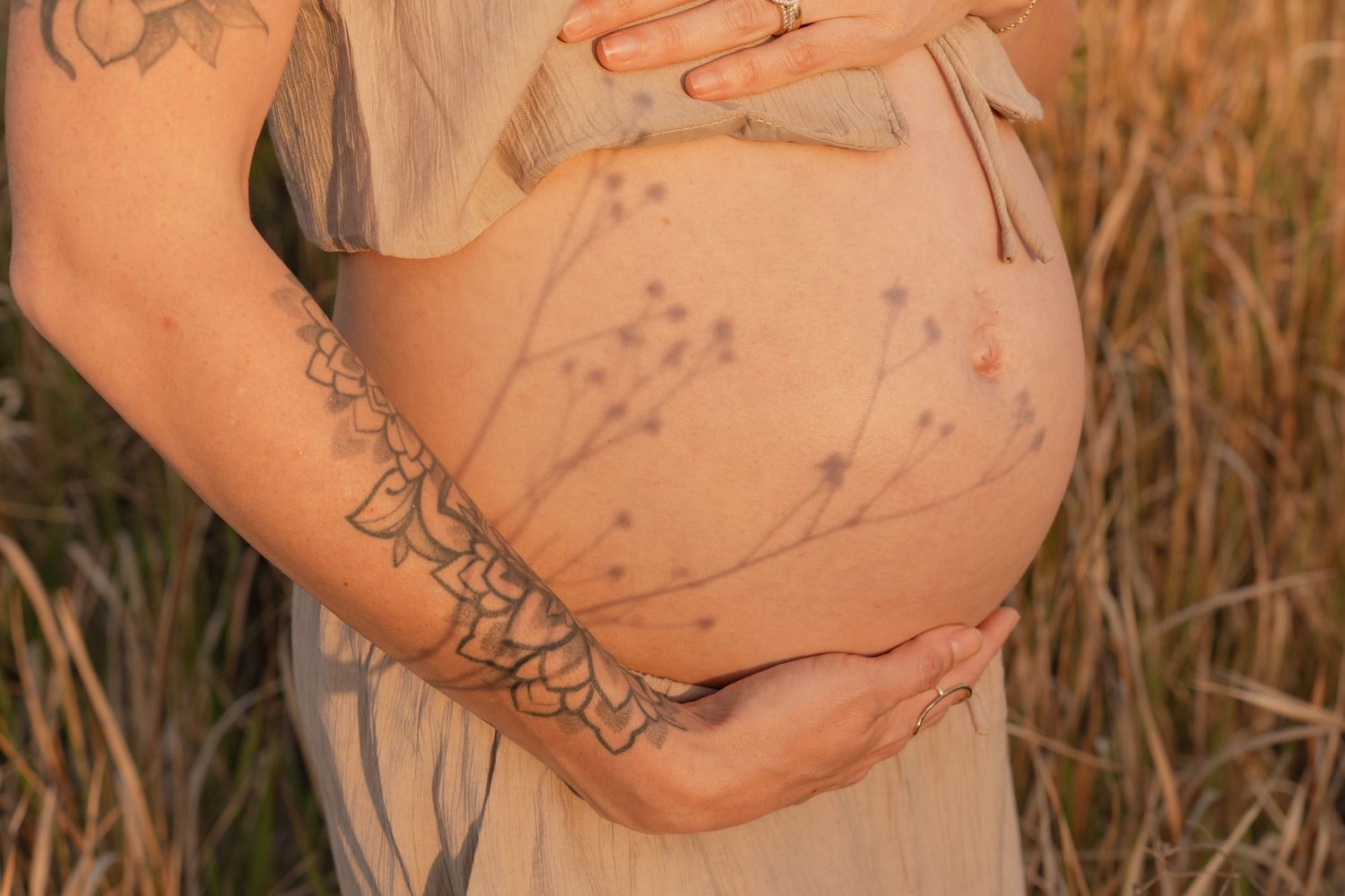 Close-up of pregnant belly with tattoo and wildflower shadows in warm golden light.