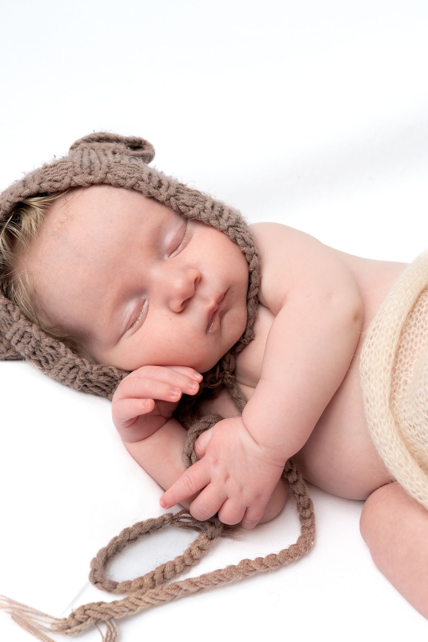 Sleeping newborn baby wearing a brown knitted bear hat with tie strings on white background.