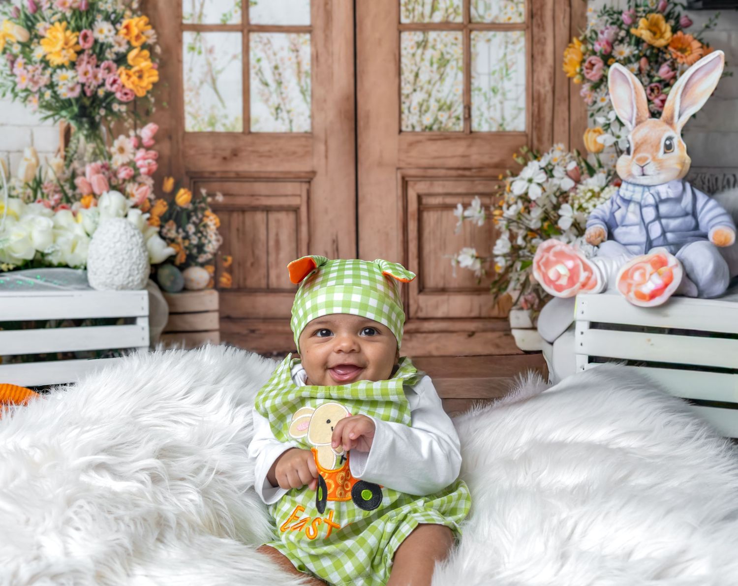 Smiling baby in green gingham outfit sits on white fur rug with Easter bunny decor and floral backdrop.