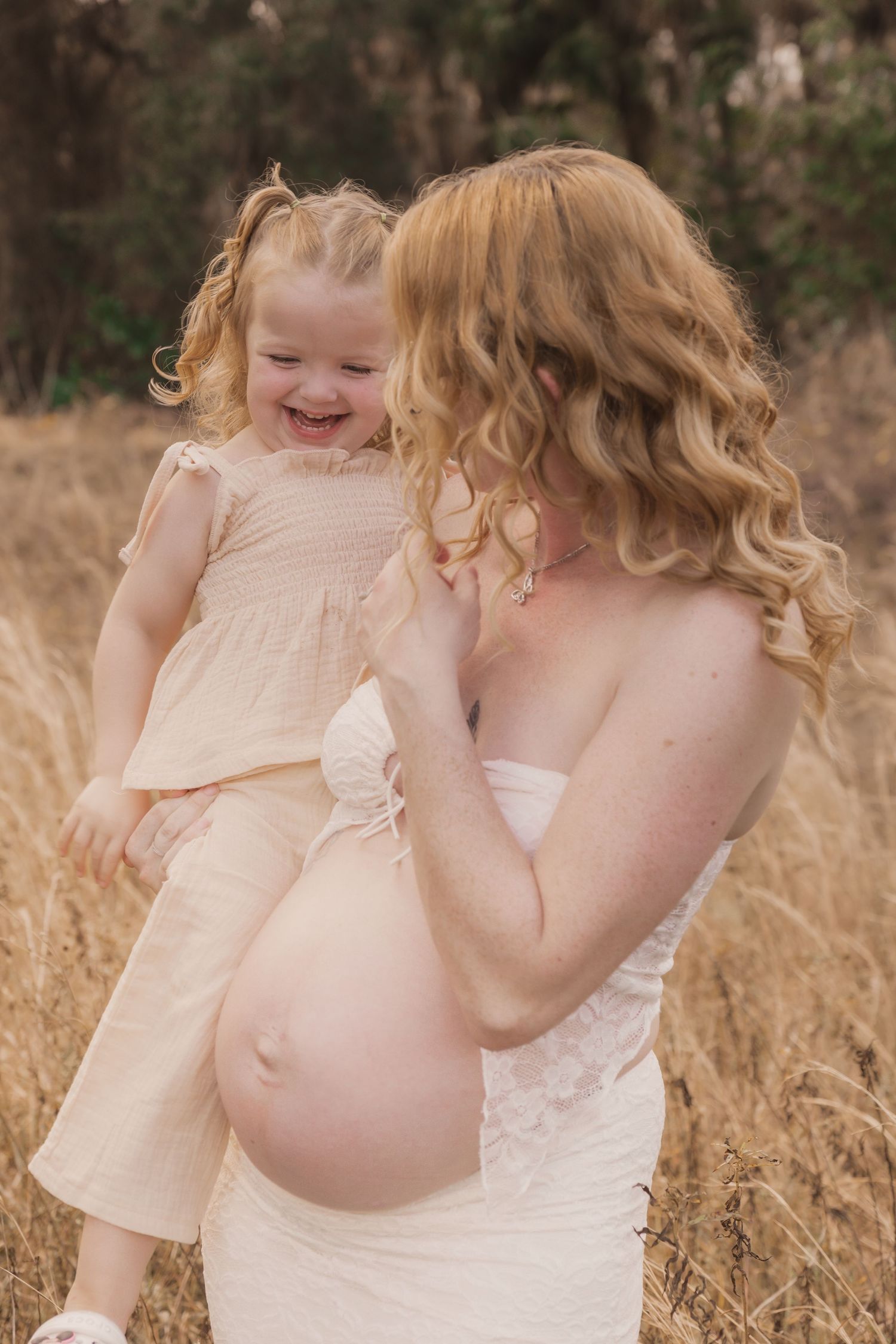 Pregnant woman in white dress holding laughing toddler girl in field of golden grass during maternity photo session.