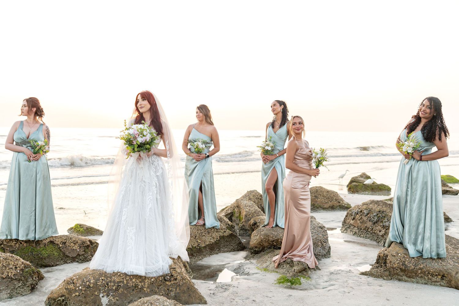 Bride in white gown with five bridesmaids in sage and blush dresses on rocky beach at sunset.
