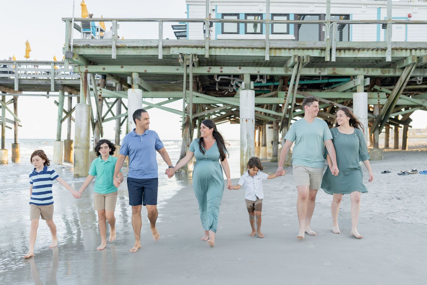 Happy family of seven walking hand-in-hand on a sunny beach near a pier.