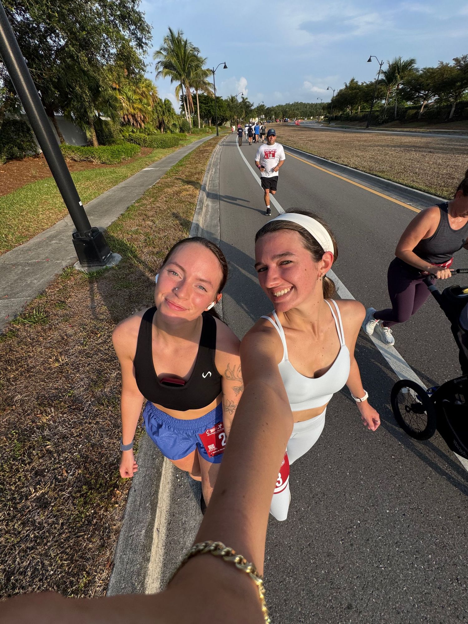 Two smiling women taking a selfie while running together on a palm-lined road.