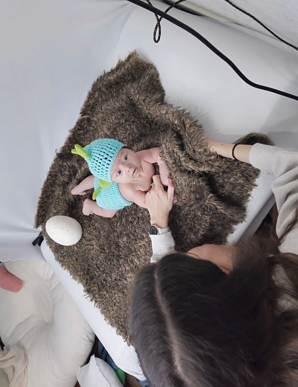 Photographer carefully positions newborn baby wearing teal knit hat on fur blanket during studio session.