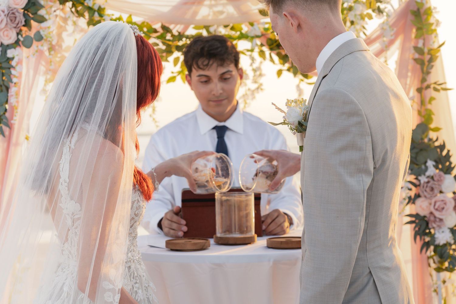 Officiant performs sand ceremony for bride and groom under floral arch at an outdoor wedding ceremony.