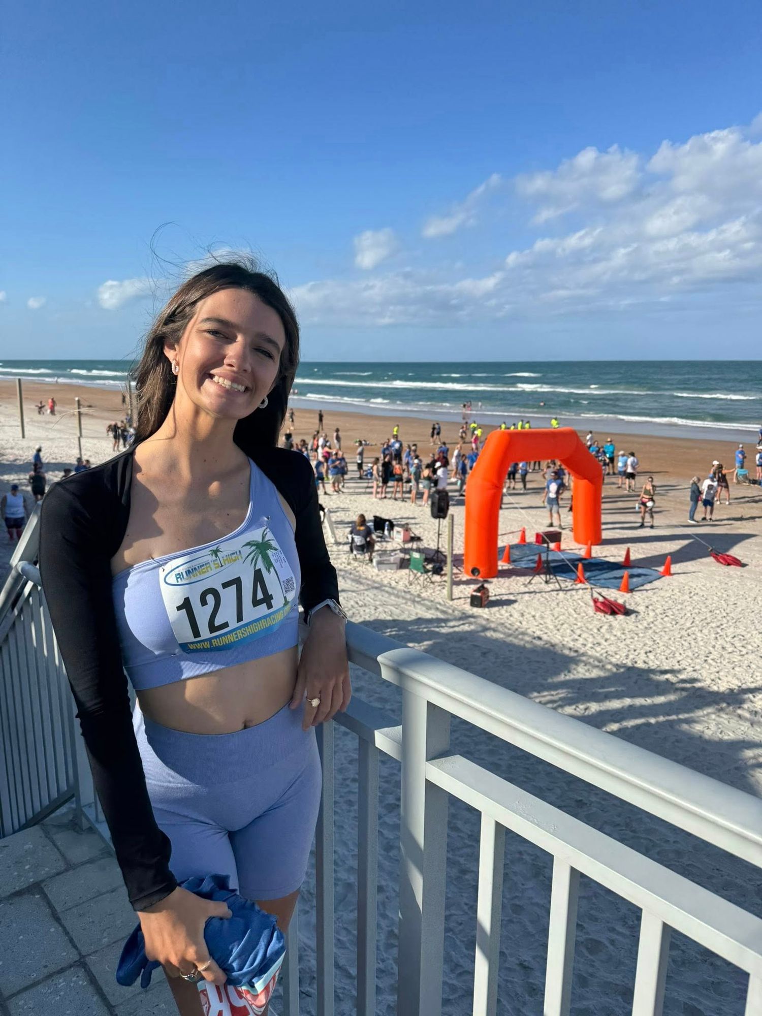 Smiling woman with race bib 1274 stands at beach triathlon finish line on sunny day.