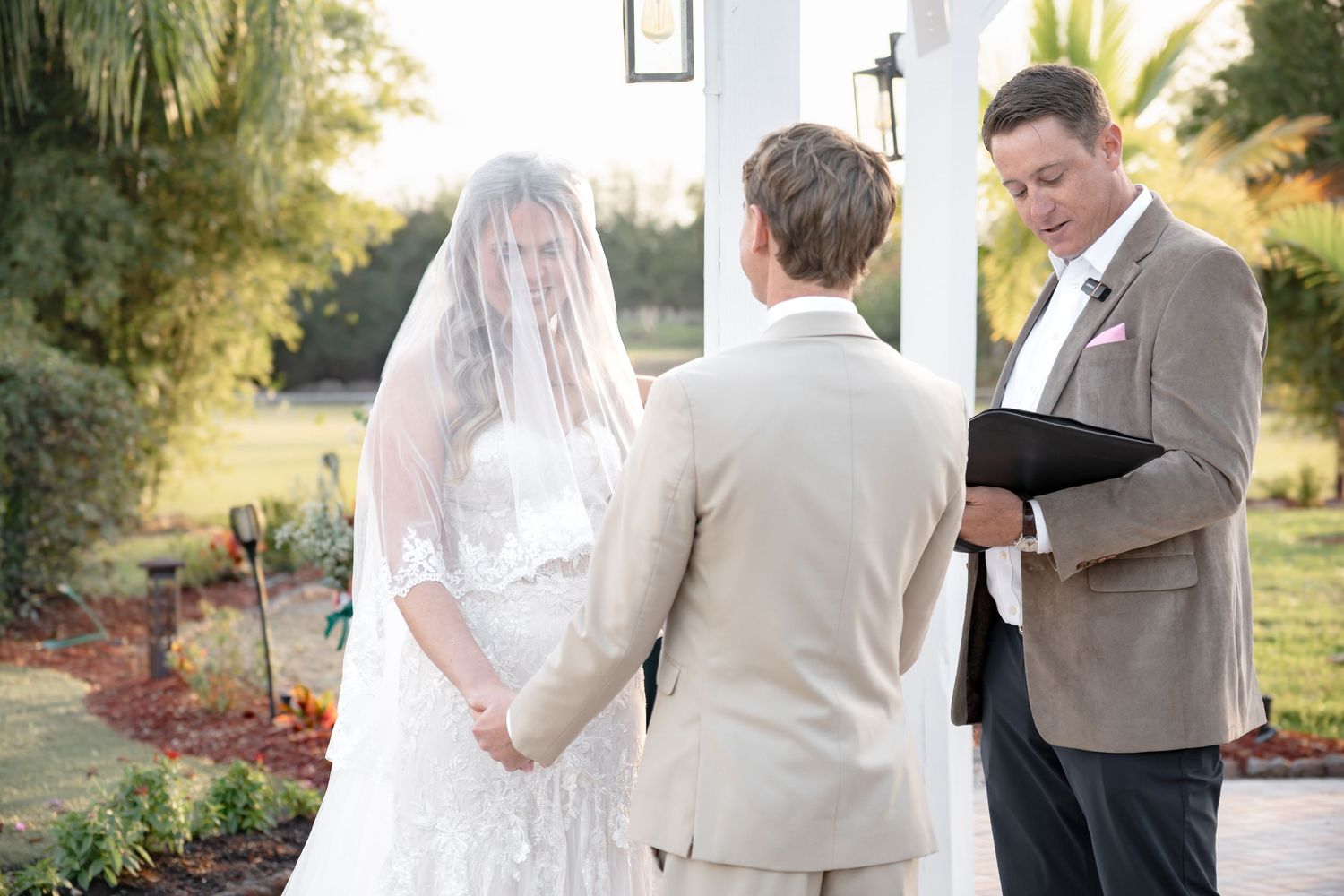 Bride and groom hold hands during an outdoor wedding ceremony while an officiant reads from a book.