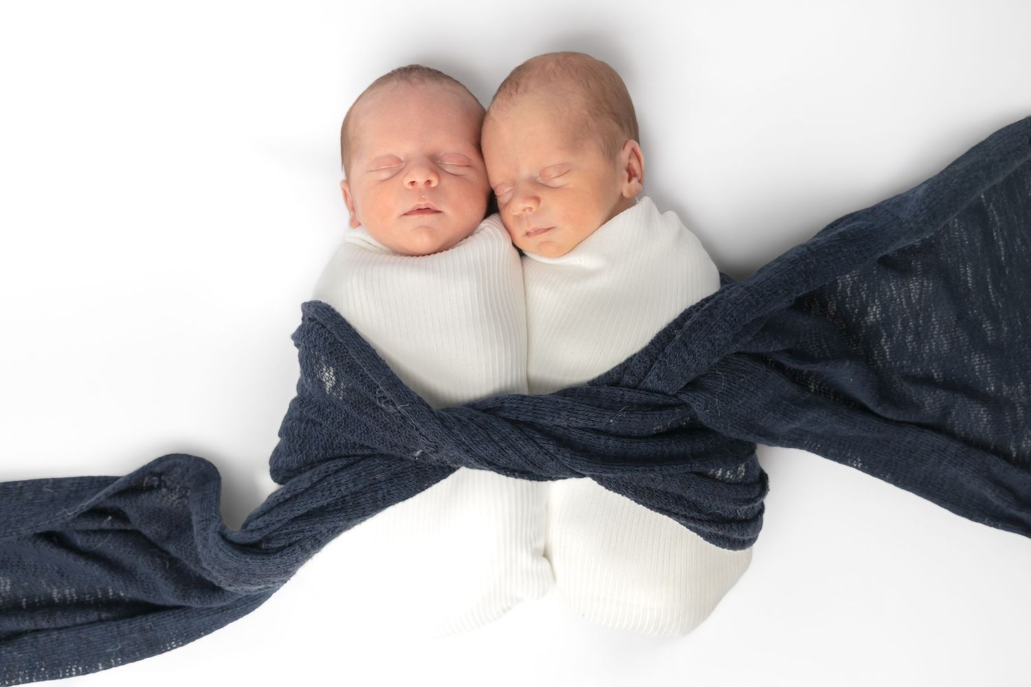 Two sleeping newborn twins swaddled together in white and navy blue blankets on a white background.