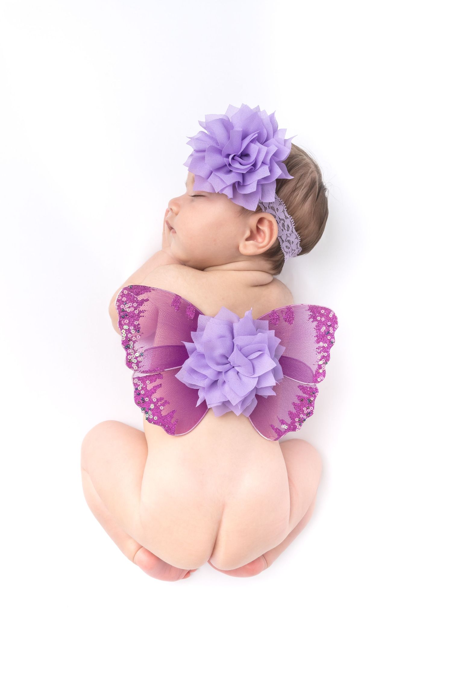 Sleeping newborn baby girl with purple butterfly wings and floral headband on white background.