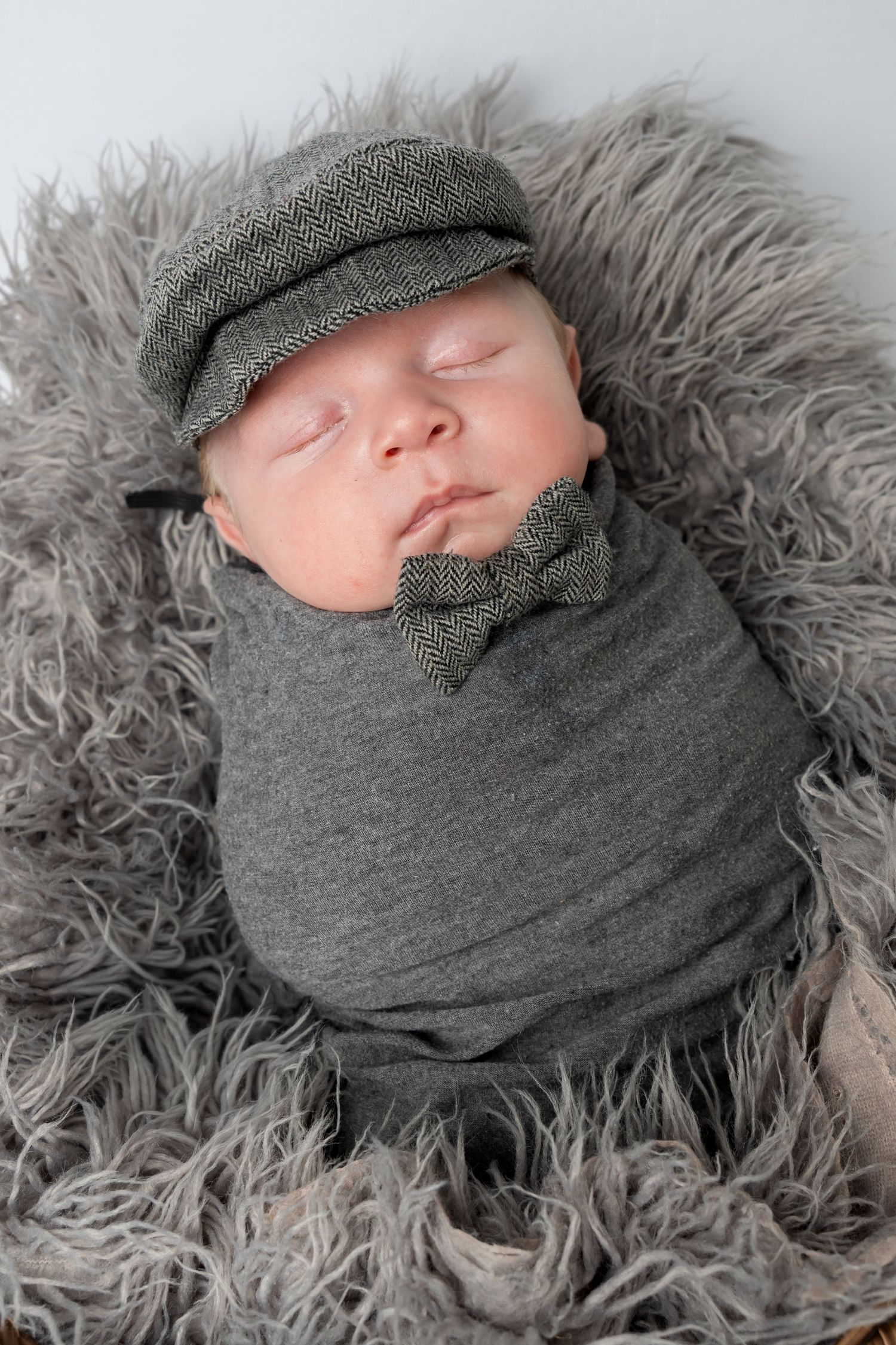 Newborn baby swaddled in gray wrap, wearing a herringbone flat cap and bow tie, sleeping in a fluffy gray basket.
