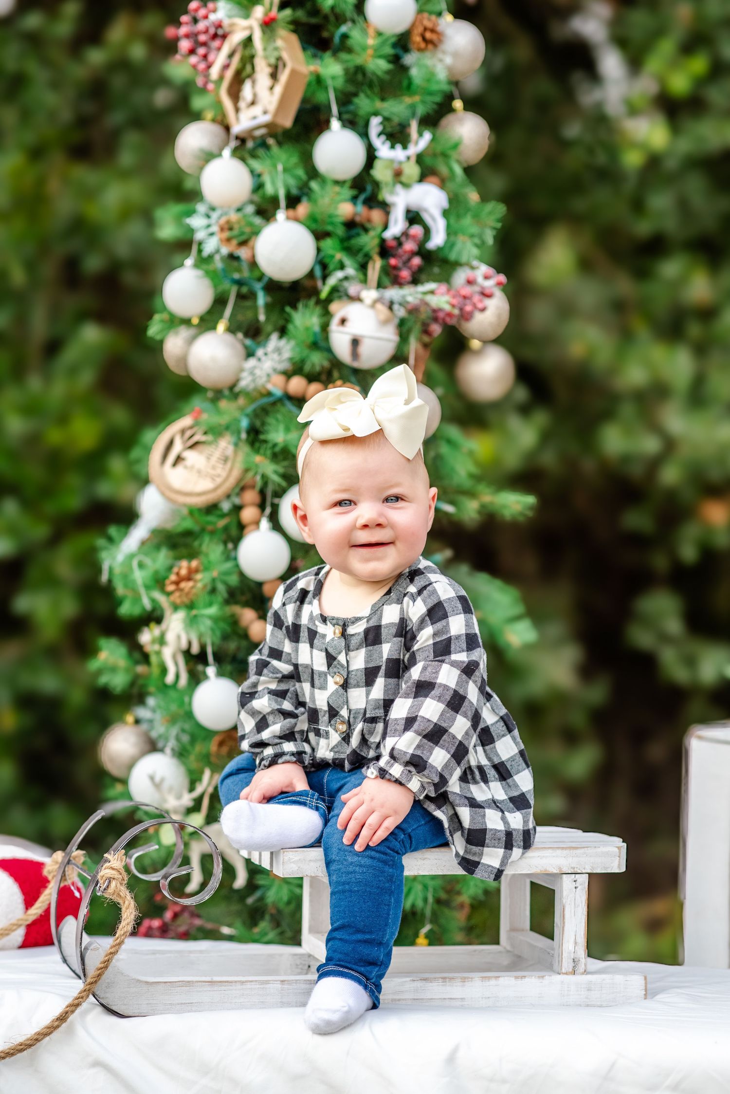Smiling baby girl in plaid shirt and bow headband sitting on white bench by decorated Christmas tree.