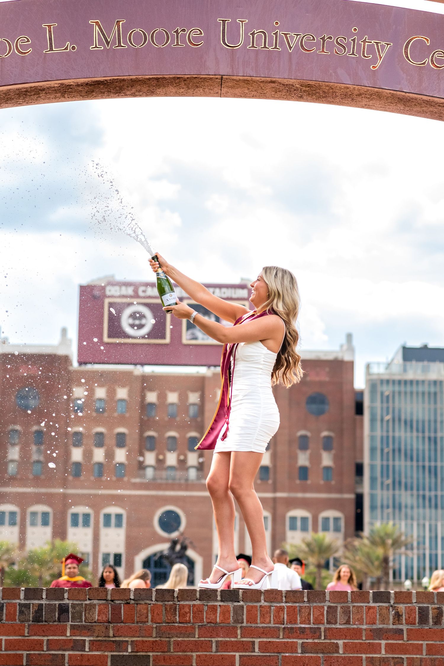 Graduate in white dress pops champagne under Moore University Center arch at Florida State University.
