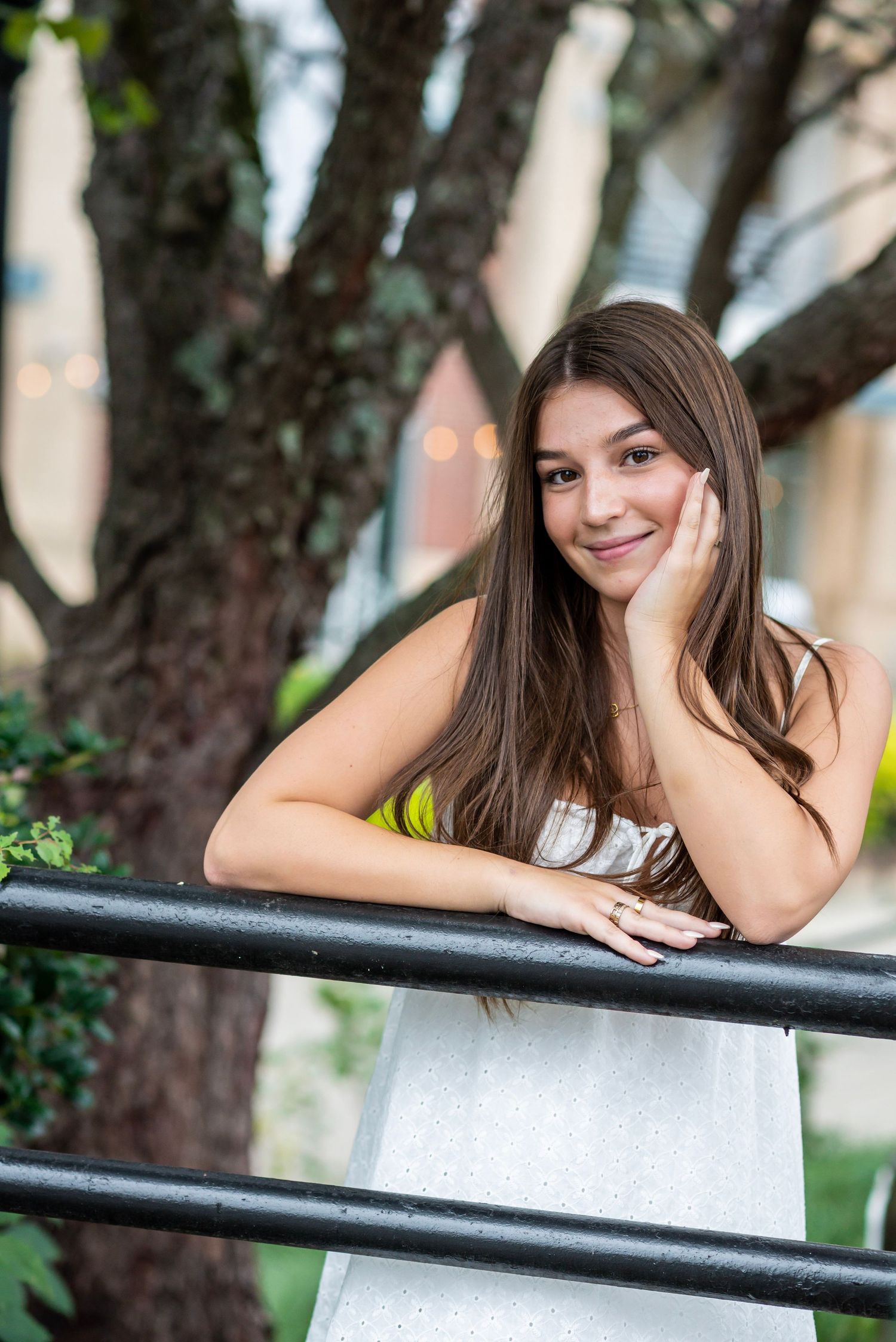 Young woman with long brown hair smiling outdoors, leaning on a black railing in a white dress.