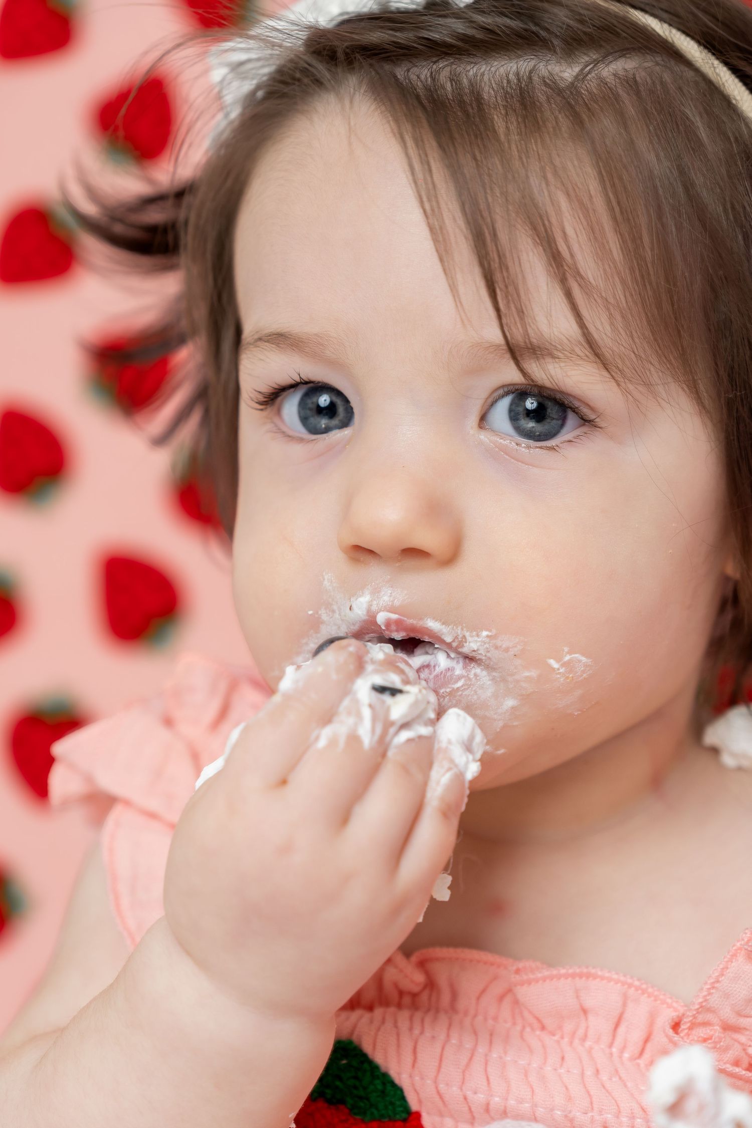 A baby girl with blue eyes eating cake with frosting on her face, wearing a pink dress with strawberry background.