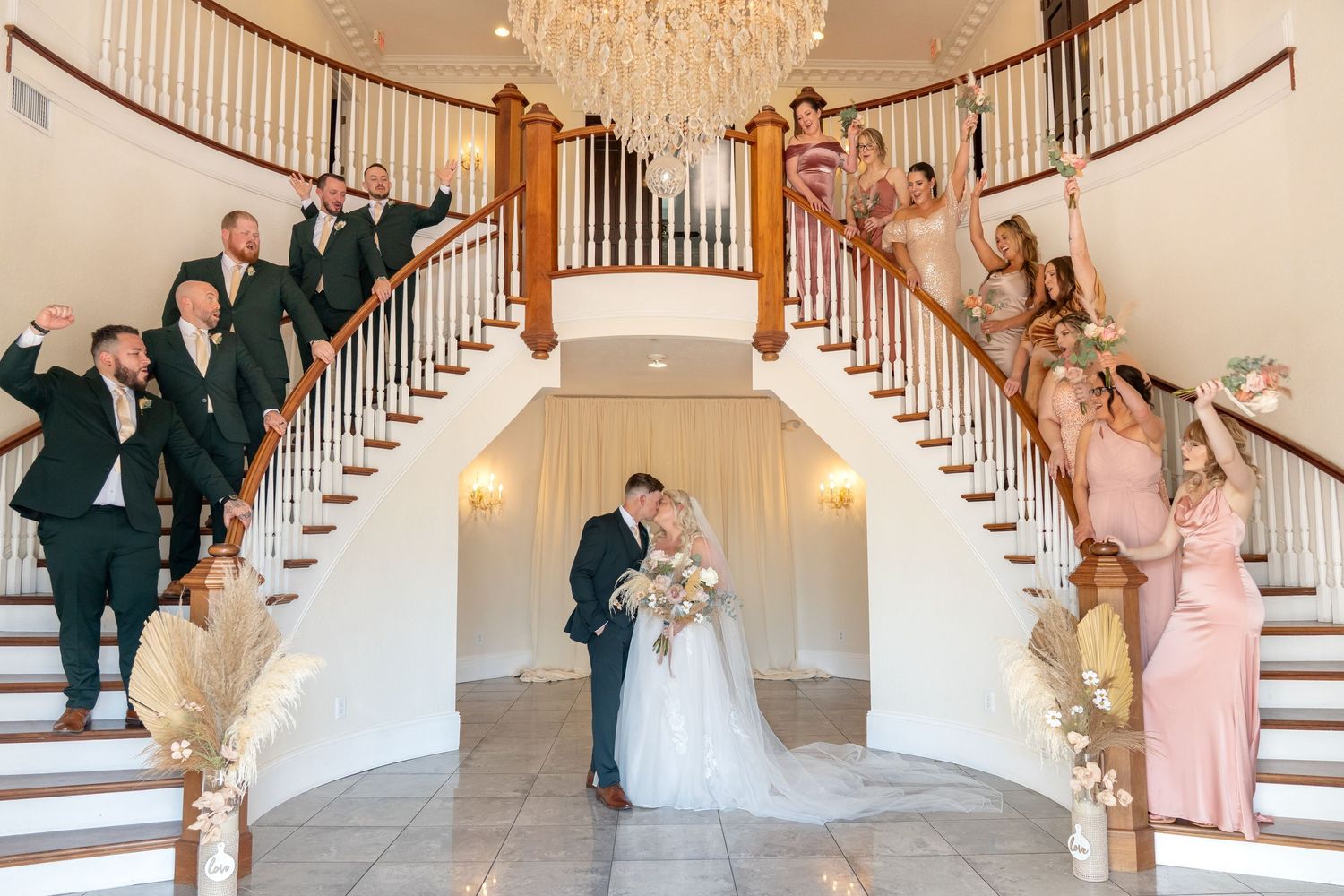 Wedding party celebrating on grand dual staircase as bride and groom share a kiss under a chandelier.