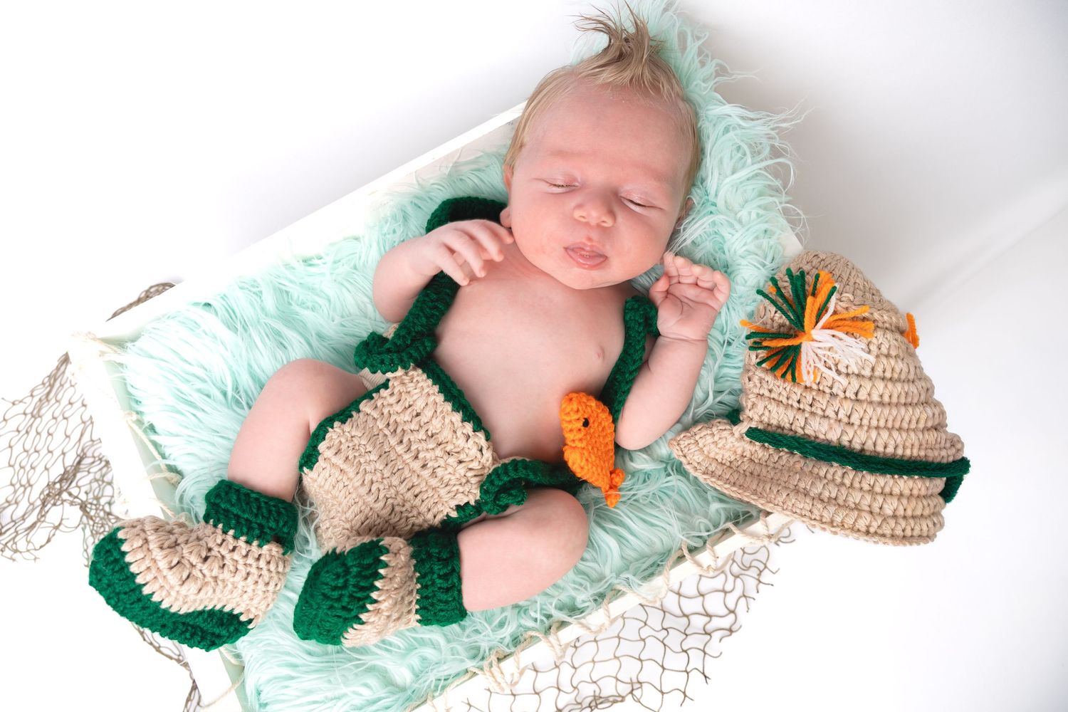 Newborn baby in crochet fishing outfit with hat, booties, and orange fish, lying on mint fur in a net basket.