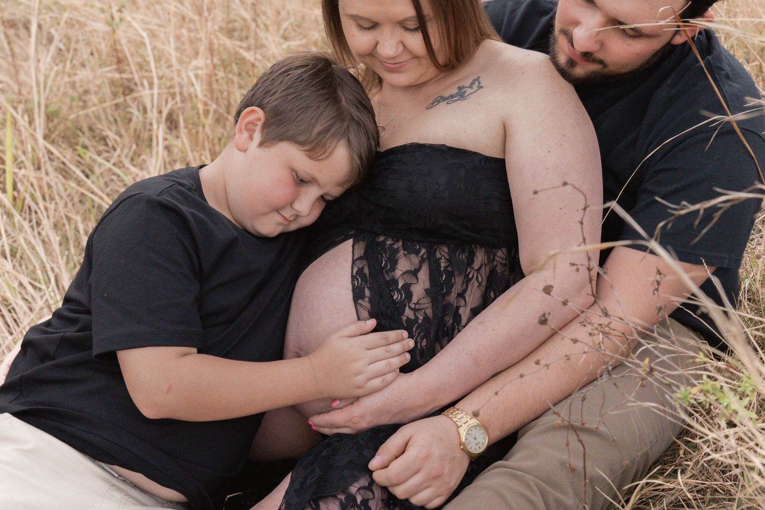 Family maternity photo session in golden field, pregnant woman in black lace dress with partner and child.