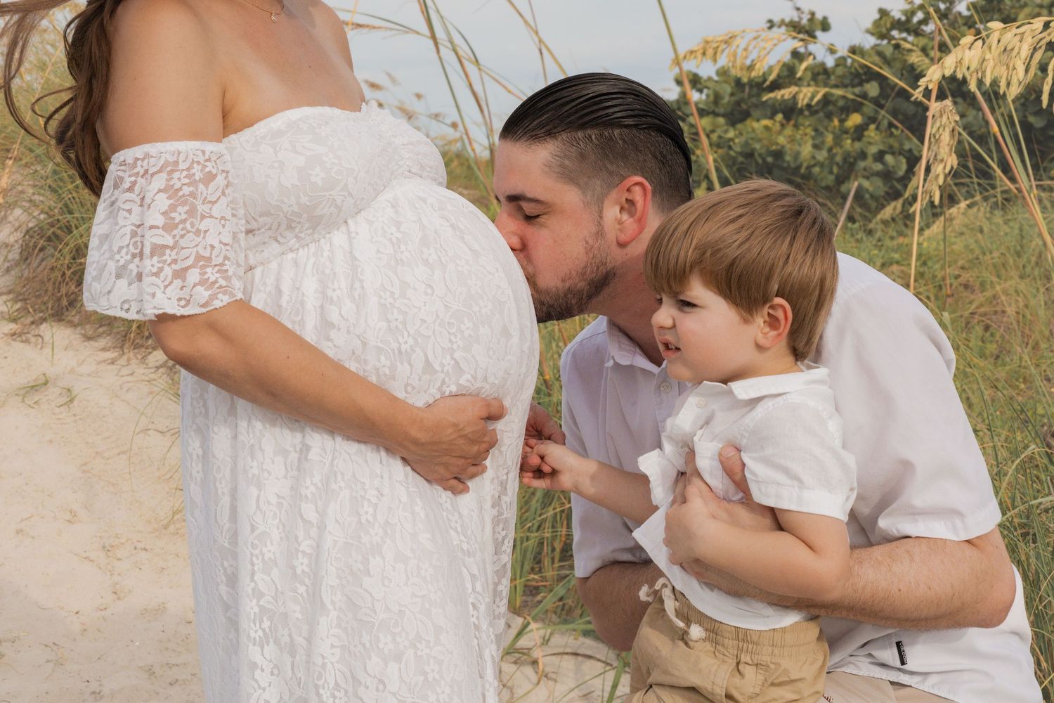 Pregnant woman in white lace dress with husband kissing belly and toddler son on sandy beach.