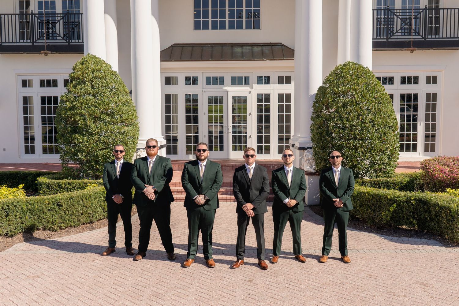 Six groomsmen in matching dark green suits pose confidently outside a grand white estate venue.