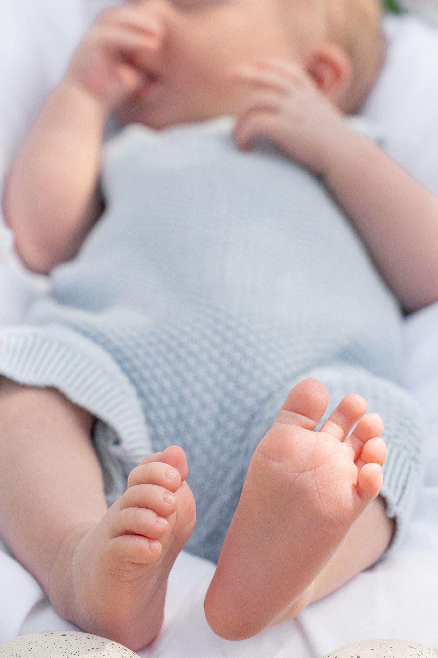 Close-up of adorable newborn baby feet and tiny toes, wearing a soft blue knit romper while lying on white blanket.