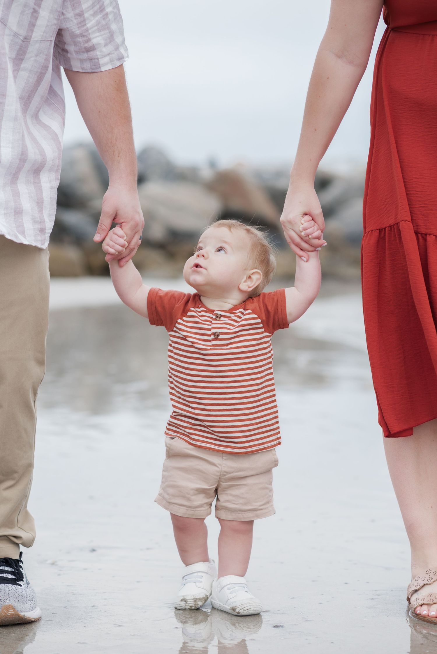 Toddler in striped shirt holds parents hands on beach, learning to walk on wet sand near rocky shore.